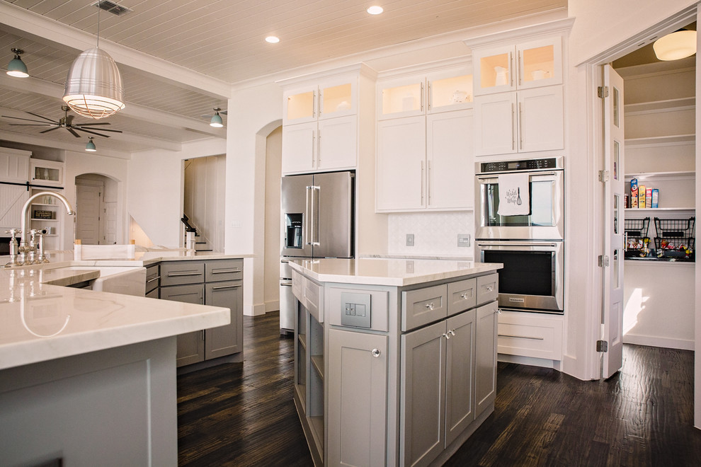 Design ideas for a large beach style l-shaped open plan kitchen in Dallas with a farmhouse sink, shaker cabinets, white cabinets, marble benchtops, white splashback, ceramic splashback, stainless steel appliances, dark hardwood floors, multiple islands and brown floor.