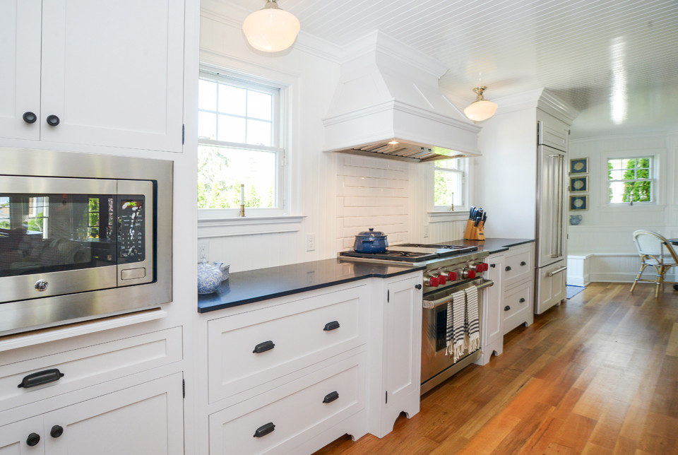 Photo of a large beach style galley open plan kitchen in Philadelphia with a farmhouse sink, shaker cabinets, white cabinets, quartz benchtops, white splashback, porcelain splashback, stainless steel appliances, medium hardwood floors and with island.
