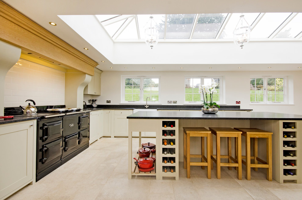 Photo of a country l-shaped kitchen in London with a farmhouse sink, shaker cabinets, beige cabinets, white splashback, subway tile splashback, black appliances and with island.