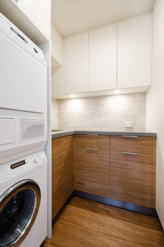 Inspiration for a small contemporary u-shaped utility room in Melbourne with flat-panel cabinets, medium wood cabinets, quartz benchtops, beige walls, plywood floors, a stacked washer and dryer and an undermount sink.