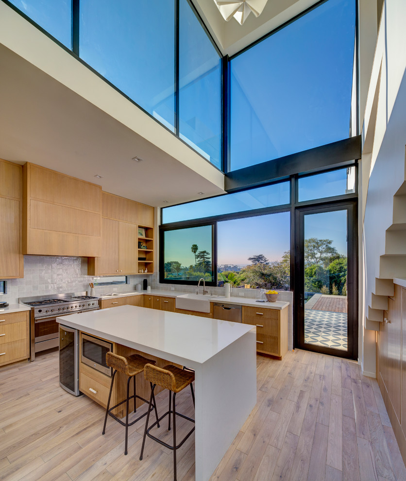 Inspiration for a large contemporary u-shaped open plan kitchen in Los Angeles with a farmhouse sink, recessed-panel cabinets, medium wood cabinets, quartz benchtops, stainless steel appliances, with island, white benchtop, light hardwood floors, beige floor and grey splashback.