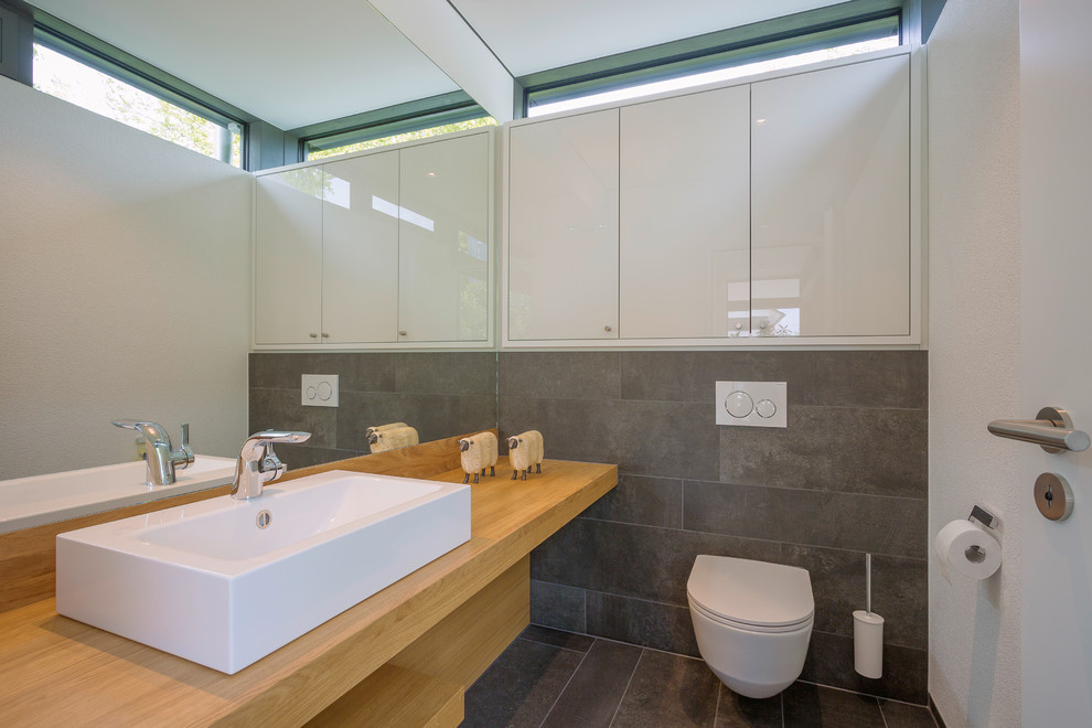 Large contemporary powder room in Other with a wall-mount toilet, gray tile, white walls, wood benchtops, grey floor, brown benchtops, flat-panel cabinets, white cabinets, ceramic tile, ceramic floors and a vessel sink.