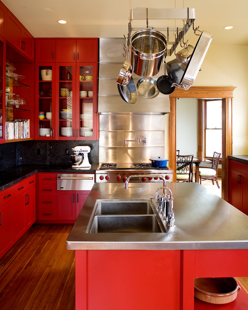 Design ideas for a transitional l-shaped kitchen in Austin with a double-bowl sink, glass-front cabinets, red cabinets, stainless steel benchtops, black splashback, stainless steel appliances, medium hardwood floors and with island.
