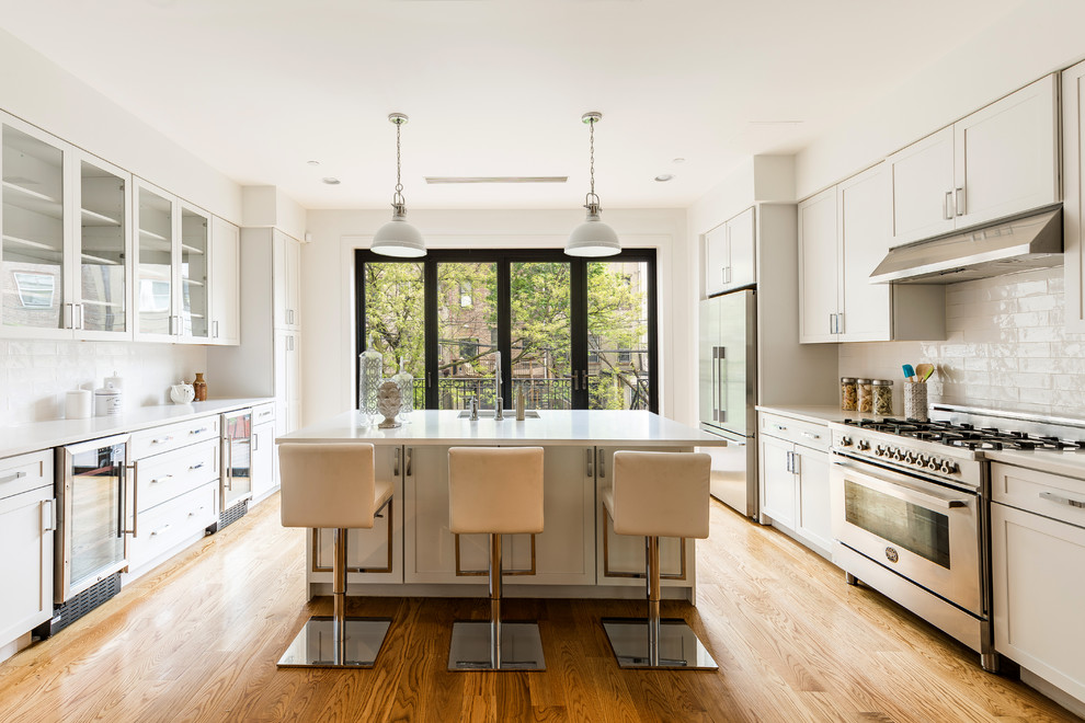 Transitional galley medium tone wood floor and brown floor kitchen photo in New York with an undermount sink, shaker cabinets, white cabinets, white backsplash, stainless steel appliances, an island and white countertops