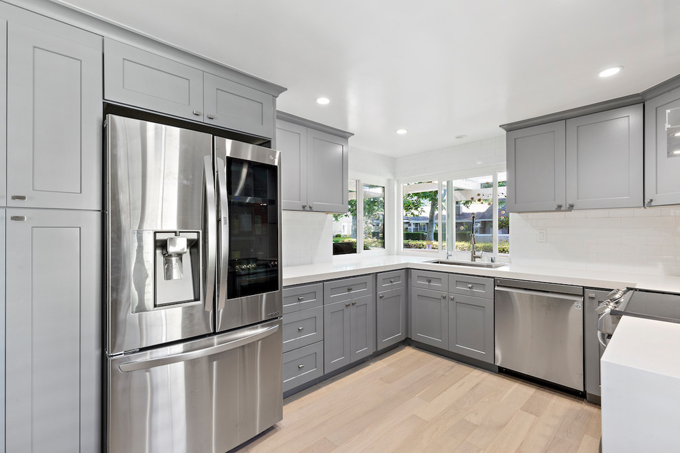 Example of a mid-sized transitional u-shaped light wood floor and beige floor enclosed kitchen design with an undermount sink, shaker cabinets, gray cabinets, solid surface countertops, white backsplash, subway tile backsplash, stainless steel appliances and white countertops