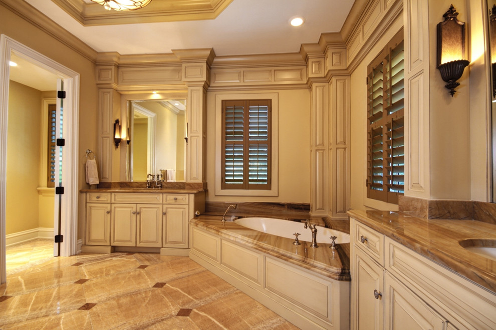 Large traditional master bathroom in Miami with raised-panel cabinets, beige cabinets, a corner tub, a one-piece toilet, beige walls, limestone floors, an undermount sink, onyx benchtops and beige floor.
