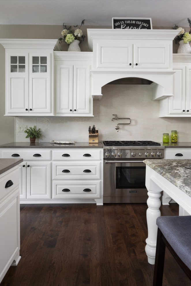 Photo of a large kitchen in Milwaukee with a farmhouse sink, raised-panel cabinets, white cabinets, granite benchtops, white splashback, ceramic splashback, stainless steel appliances, medium hardwood floors, with island, brown floor and grey benchtop.