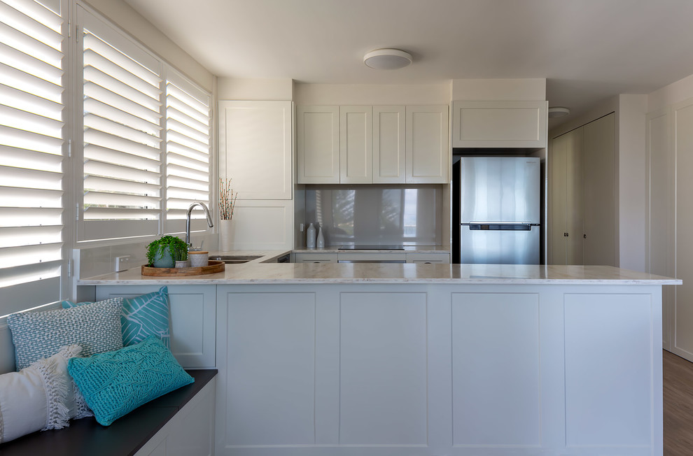 Mid-sized contemporary u-shaped open plan kitchen in Sunshine Coast with an undermount sink, shaker cabinets, white cabinets, solid surface benchtops, metallic splashback, mirror splashback, vinyl floors, a peninsula, multi-coloured floor and white benchtop.