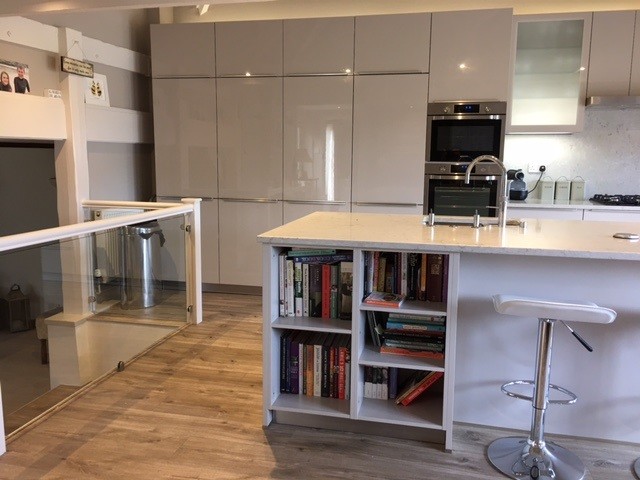 Photo of a mid-sized contemporary single-wall open plan kitchen in Oxfordshire with an integrated sink, flat-panel cabinets, beige cabinets, solid surface benchtops, grey splashback, stone slab splashback, panelled appliances, laminate floors and with island.