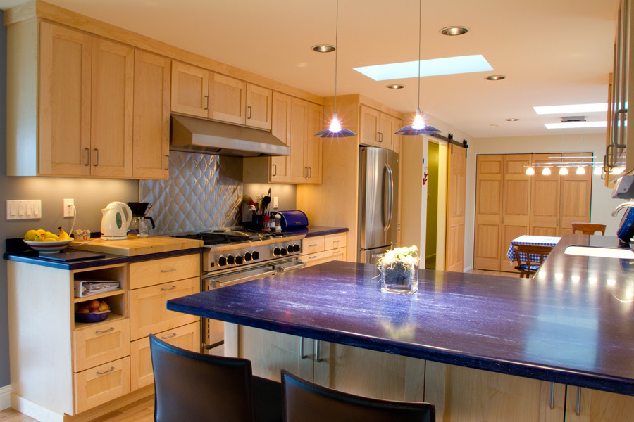 This is an example of a mid-sized eclectic l-shaped open plan kitchen in Seattle with an undermount sink, shaker cabinets, light wood cabinets, solid surface benchtops, blue splashback, stainless steel appliances, light hardwood floors and a peninsula.