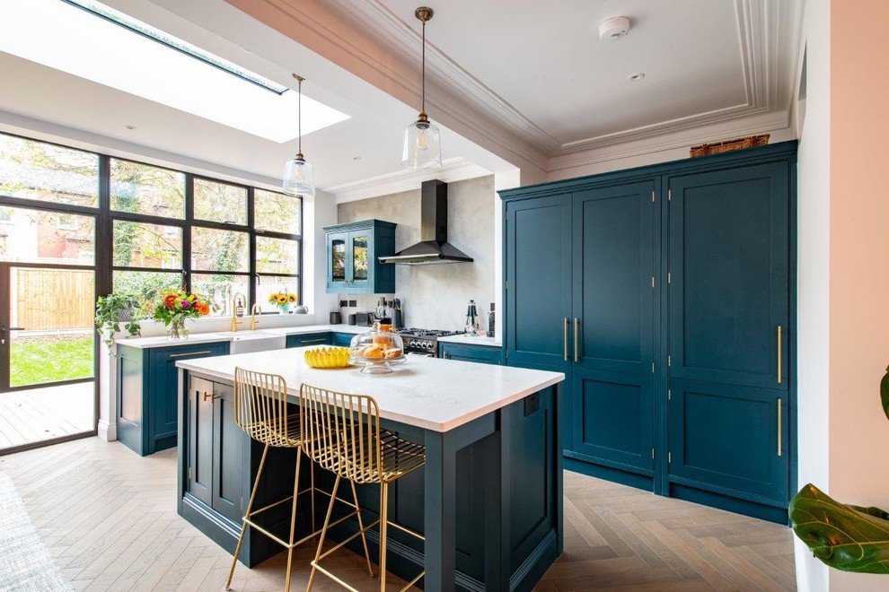Transitional l-shaped kitchen in Kent with a farmhouse sink, shaker cabinets, turquoise cabinets, grey splashback, light hardwood floors, with island, white benchtop and beige floor.