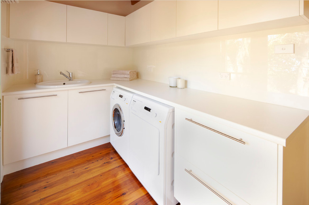 This is an example of a transitional l-shaped laundry room in Sydney with a drop-in sink, flat-panel cabinets, white cabinets, white walls, light hardwood floors and a side-by-side washer and dryer.