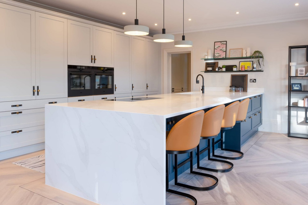 This is an example of a large transitional single-wall open plan kitchen in Hertfordshire with a single-bowl sink, recessed-panel cabinets, grey cabinets, quartzite benchtops, black appliances, light hardwood floors, with island, brown floor and white benchtop.