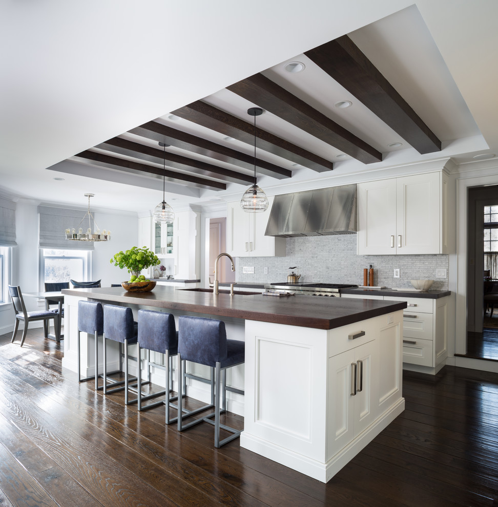 This is an example of a transitional galley eat-in kitchen in New York with an undermount sink, recessed-panel cabinets, white cabinets, wood benchtops, stone tile splashback, stainless steel appliances, dark hardwood floors, with island and grey splashback.