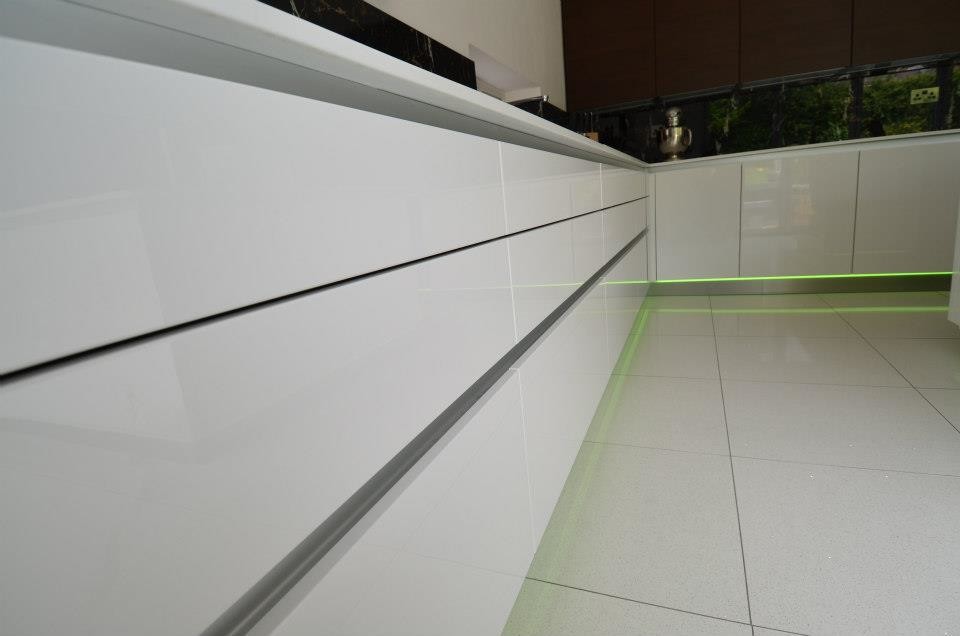 Expansive contemporary l-shaped eat-in kitchen in West Midlands with a single-bowl sink, flat-panel cabinets, white cabinets, solid surface benchtops, multi-coloured splashback, glass tile splashback, black appliances, porcelain floors and with island.
