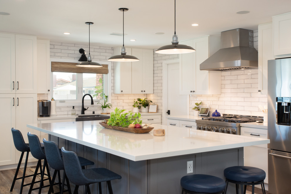 Example of a mid-sized transitional l-shaped brown floor open concept kitchen design in Orange County with shaker cabinets, white cabinets, white backsplash, stainless steel appliances, an island, white countertops and quartz countertops