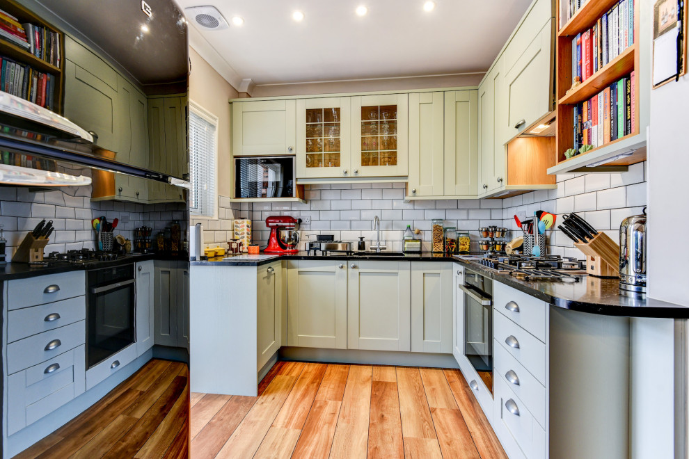 Inspiration for a medium sized traditional grey and white l-shaped kitchen/diner in Sussex with a submerged sink, shaker cabinets, green cabinets, quartz worktops, white splashback, ceramic splashback, black appliances, light hardwood flooring, no island, brown floors, black worktops and feature lighting.