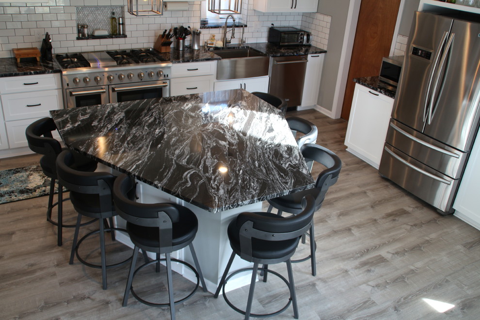 Photo of a mid-sized beach style l-shaped eat-in kitchen in Detroit with a farmhouse sink, shaker cabinets, white cabinets, granite benchtops, white splashback, ceramic splashback, stainless steel appliances, vinyl floors, with island, grey floor and black benchtop.