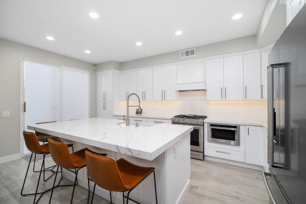 Inspiration for a mid-sized transitional laminate floor and gray floor kitchen remodel in Orange County with a farmhouse sink, shaker cabinets, white cabinets, white backsplash, subway tile backsplash, stainless steel appliances, an island and white countertops