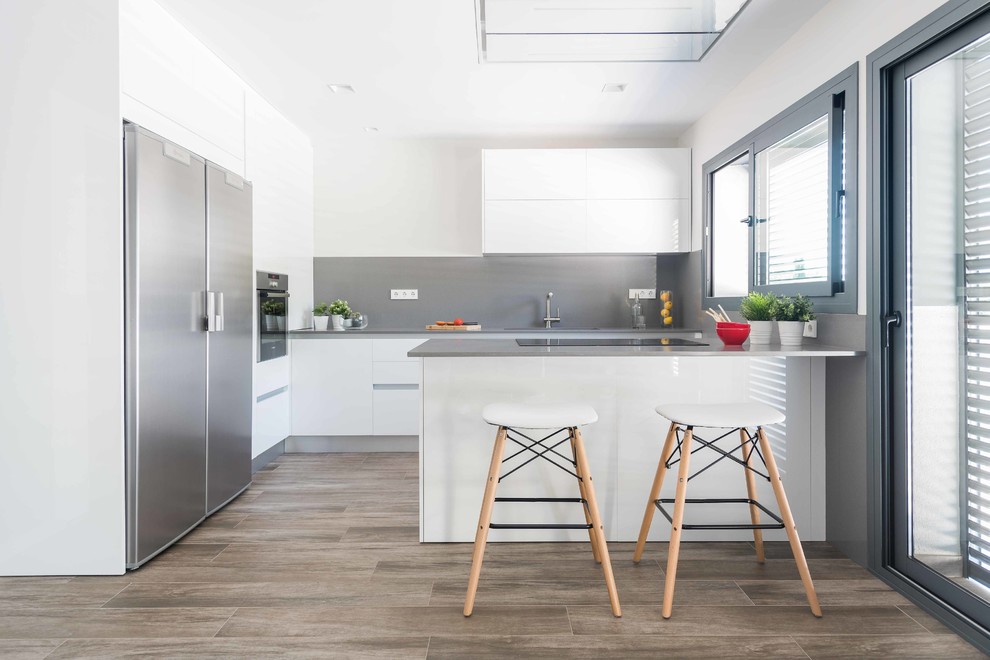 This is an example of a large contemporary u-shaped kitchen in Barcelona with a double-bowl sink, flat-panel cabinets, white cabinets, grey splashback, stainless steel appliances, ceramic floors, brown floor, grey benchtop and a peninsula.