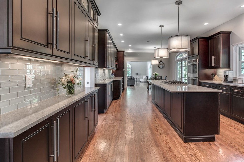 Inspiration for a large transitional galley light wood floor and brown floor eat-in kitchen remodel in Raleigh with an undermount sink, raised-panel cabinets, dark wood cabinets, quartz countertops, gray backsplash, glass tile backsplash, stainless steel appliances, an island and gray countertops