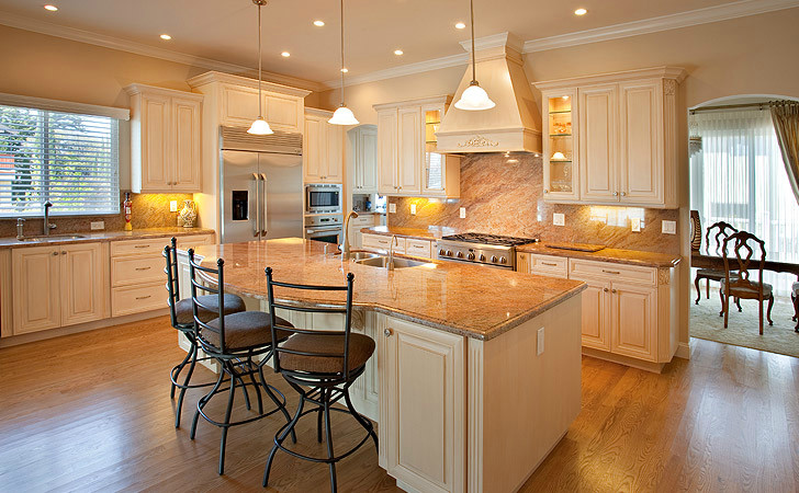 Inspiration for a large coastal l-shaped medium tone wood floor eat-in kitchen remodel with a drop-in sink, white cabinets, stainless steel appliances, beige backsplash, an island, raised-panel cabinets, granite countertops and stone slab backsplash