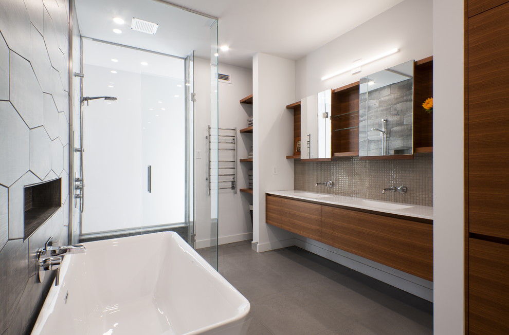 This is an example of a contemporary master bathroom in Montreal with flat-panel cabinets, medium wood cabinets, a freestanding tub, gray tile, mosaic tile, white walls, an integrated sink, grey floor and a hinged shower door.