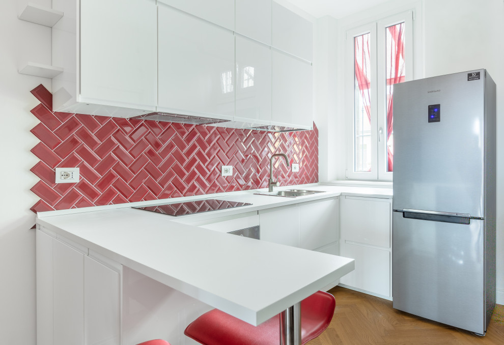 Photo of a mid-sized contemporary u-shaped open plan kitchen in Milan with a drop-in sink, flat-panel cabinets, white cabinets, red splashback, subway tile splashback, stainless steel appliances, a peninsula, brown floor, white benchtop and light hardwood floors.