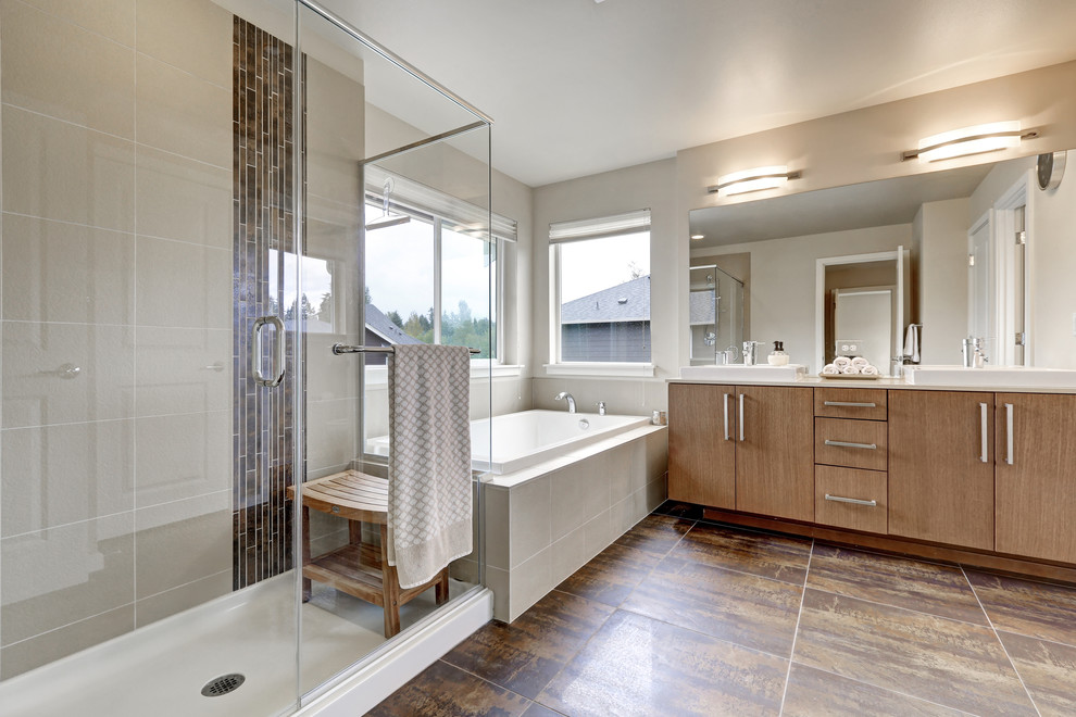 Photo of a mid-sized modern master bathroom in New York with flat-panel cabinets, medium wood cabinets, a drop-in tub, a corner shower, gray tile, ceramic tile, grey walls, ceramic floors, a trough sink, brown floor, a hinged shower door and solid surface benchtops.