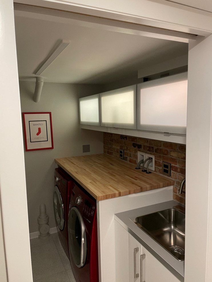Dedicated laundry room - mid-sized 1950s galley concrete floor and white floor dedicated laundry room idea in Chicago with a drop-in sink, glass-front cabinets, white cabinets, wood countertops, brick backsplash and a side-by-side washer/dryer