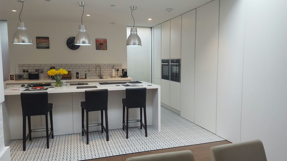 Mid-sized modern eat-in kitchen in London with a drop-in sink, flat-panel cabinets, white cabinets, quartzite benchtops, white splashback, brick splashback, panelled appliances, dark hardwood floors, with island, brown floor and white benchtop.