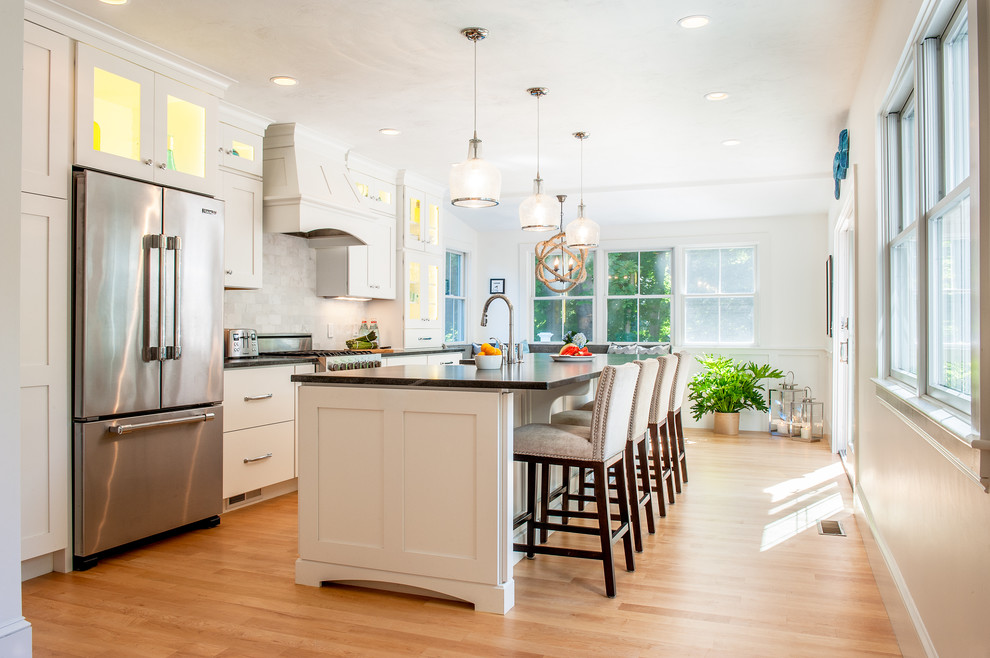 Inspiration for a beach style galley kitchen in Boston with a farmhouse sink, shaker cabinets, white cabinets, white splashback, stainless steel appliances, light hardwood floors and with island.