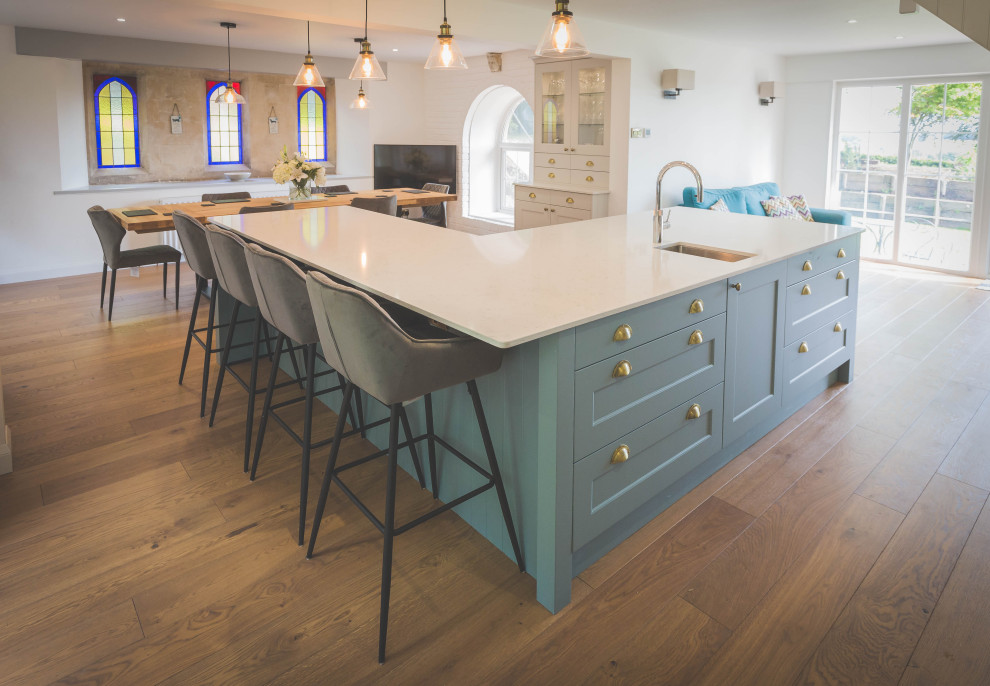 Design ideas for a large contemporary grey and teal l-shaped open plan kitchen in Devon with a belfast sink, shaker cabinets, turquoise cabinets, quartz worktops, blue splashback, glass sheet splashback, black appliances, light hardwood flooring, an island, brown floors, white worktops and feature lighting.