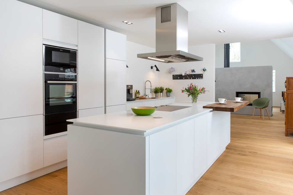 Inspiration for a large modern galley open plan kitchen in Hamburg with flat-panel cabinets, white cabinets, solid surface benchtops, white splashback, black appliances, light hardwood floors, with island, white benchtop and beige floor.