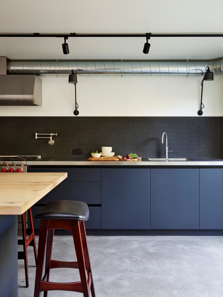 Industrial kitchen in London with an integrated sink, flat-panel cabinets, black cabinets, stainless steel benchtops, black splashback, stainless steel appliances, concrete floors, with island, grey floor and grey benchtop.