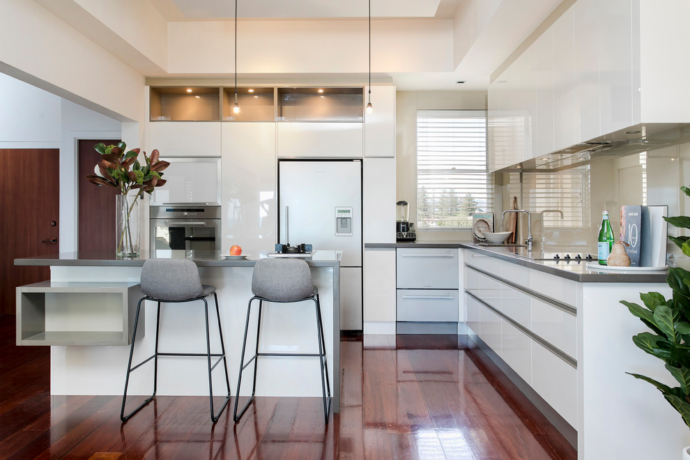 Design ideas for a contemporary l-shaped kitchen in Adelaide with an undermount sink, flat-panel cabinets, white cabinets, beige splashback, glass sheet splashback, white appliances, medium hardwood floors, with island, brown floor and grey benchtop.