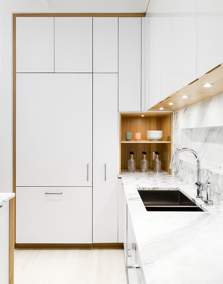 Mid-sized scandinavian l-shaped open plan kitchen in New York with flat-panel cabinets, white cabinets, panelled appliances, light hardwood floors, with island, marble benchtops, white splashback, stone slab splashback and an undermount sink.