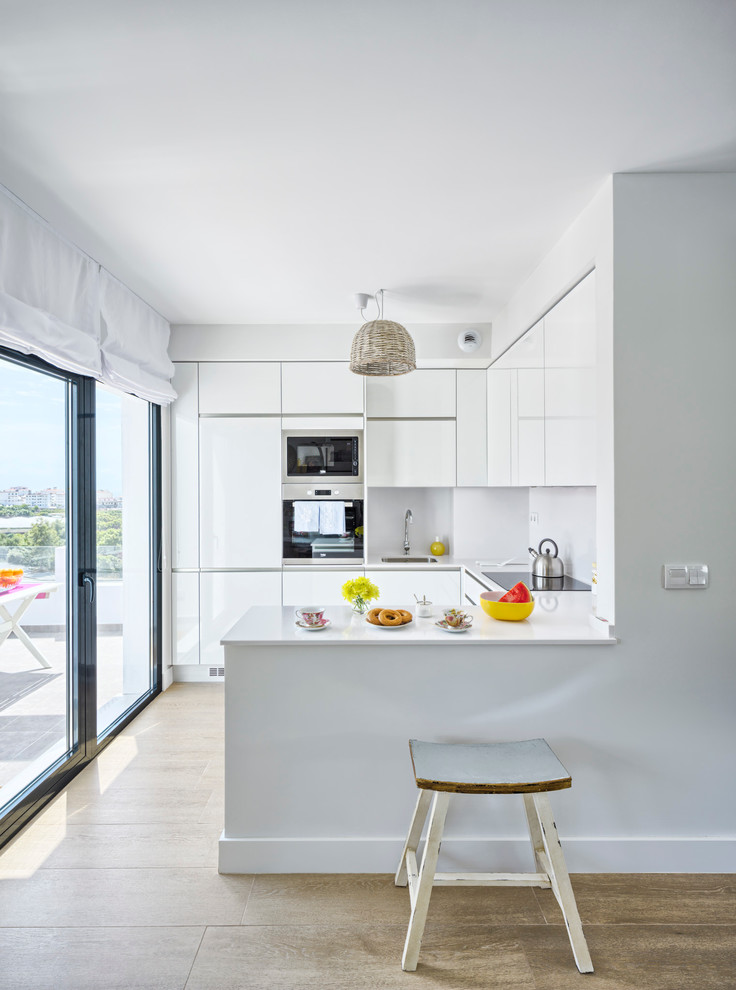 This is an example of a small beach style u-shaped open plan kitchen in Malaga with a drop-in sink, flat-panel cabinets, white cabinets, quartz benchtops, white splashback and porcelain floors.