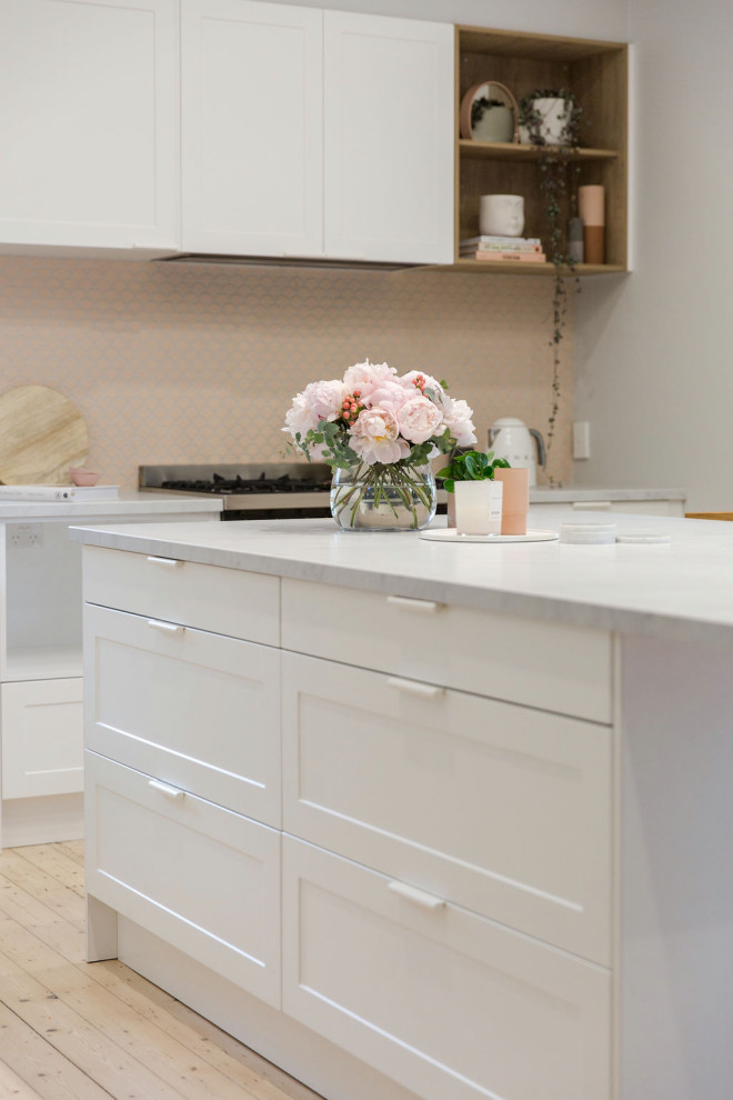 Photo of a small scandinavian l-shaped eat-in kitchen in Melbourne with an undermount sink, recessed-panel cabinets, white cabinets, quartz benchtops, pink splashback, mosaic tile splashback, black appliances, medium hardwood floors, with island and white benchtop.