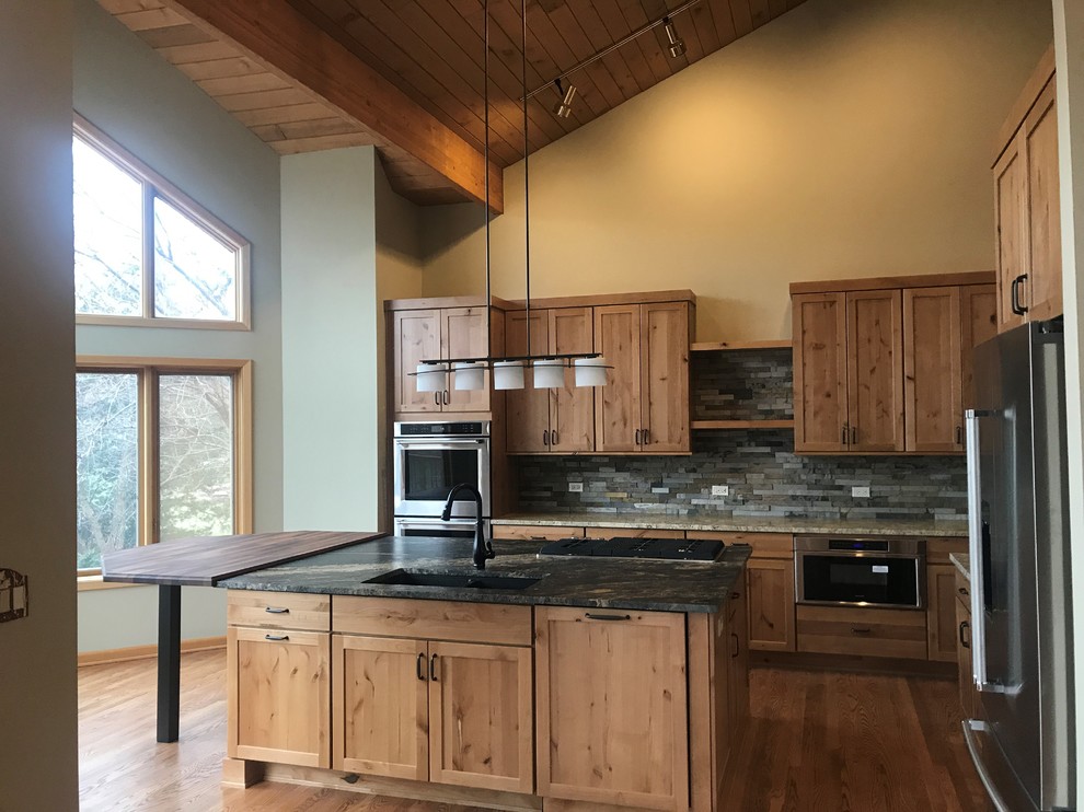 This is an example of a large country l-shaped open plan kitchen in Chicago with an undermount sink, shaker cabinets, medium wood cabinets, granite benchtops, green splashback, slate splashback, stainless steel appliances, medium hardwood floors, with island, brown floor and black benchtop.