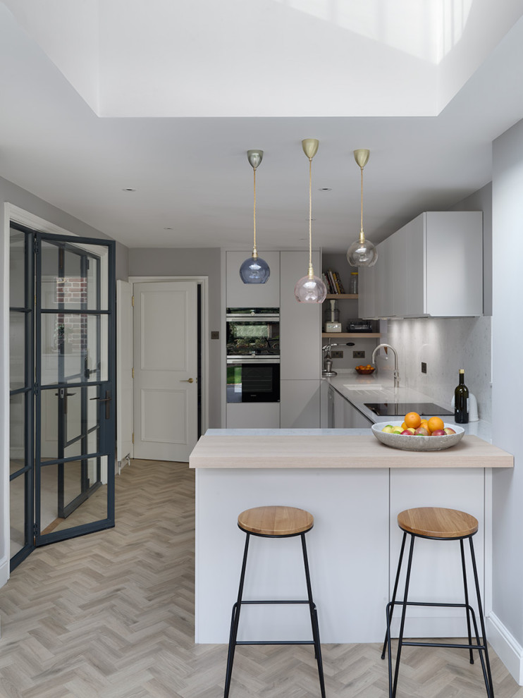Photo of a contemporary separate kitchen in London with an undermount sink, flat-panel cabinets, grey cabinets, grey splashback, black appliances, a peninsula, beige floor and grey benchtop.