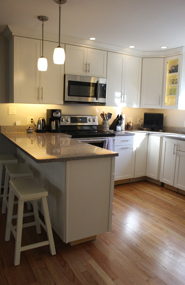 This is an example of a mid-sized transitional u-shaped eat-in kitchen in Portland Maine with a double-bowl sink, shaker cabinets, white cabinets, quartz benchtops, grey splashback, stone slab splashback, stainless steel appliances, medium hardwood floors, a peninsula and brown floor.