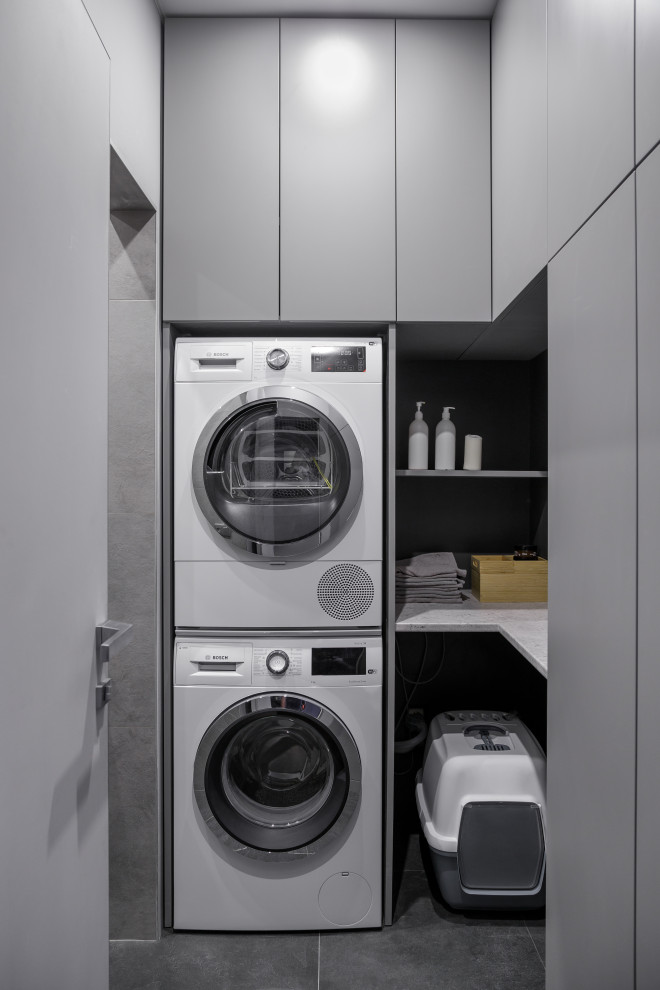 Inspiration for a small contemporary l-shaped dedicated laundry room in Saint Petersburg with flat-panel cabinets, grey cabinets, grey walls, porcelain floors, black floor, grey benchtop, a stacked washer and dryer and black splashback.