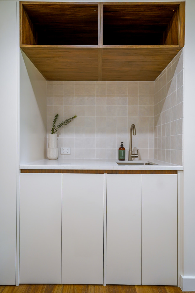 Photo of a small contemporary single-wall utility room in Sydney with a drop-in sink, flat-panel cabinets, white cabinets, granite benchtops, white walls, medium hardwood floors, a concealed washer and dryer, brown floor and white benchtop.
