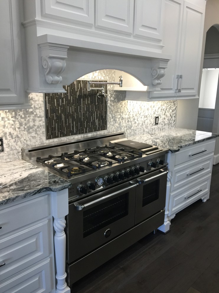 Mid-sized transitional l-shaped dark wood floor and brown floor open concept kitchen photo in Orange County with a farmhouse sink, raised-panel cabinets, white cabinets, marble countertops, metallic backsplash, metal backsplash, stainless steel appliances and an island