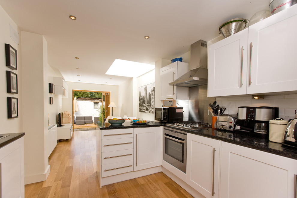 Photo of a small contemporary galley open plan kitchen in London with an integrated sink, shaker cabinets, white cabinets, granite benchtops, white splashback, ceramic splashback, stainless steel appliances, light hardwood floors, a peninsula and black benchtop.