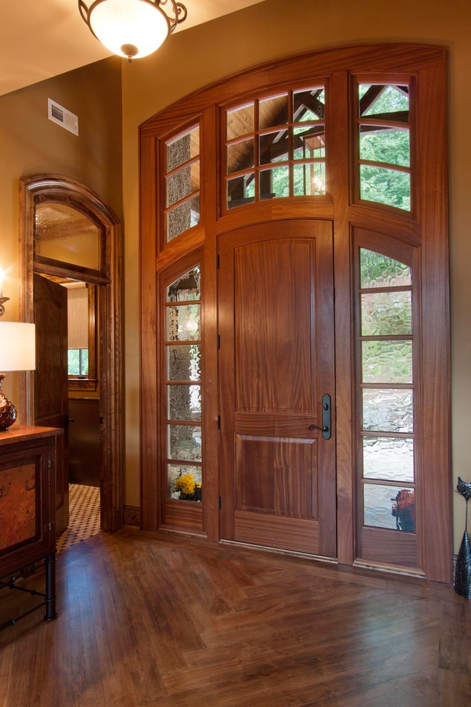 Huge mountain style brown floor and medium tone wood floor foyer photo with brown walls and a medium wood front door