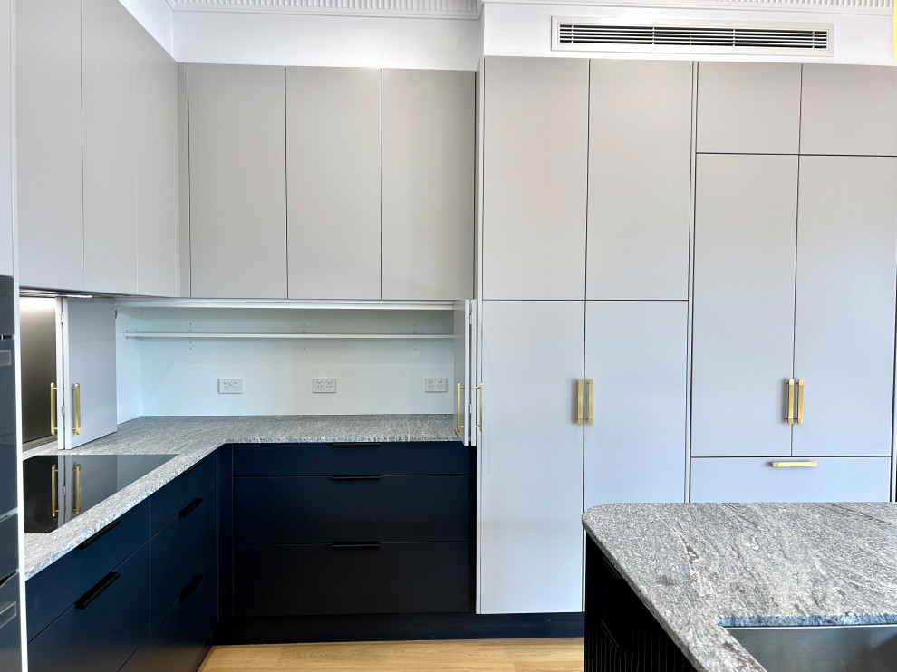 Photo of a large transitional l-shaped eat-in kitchen in Sydney with an undermount sink, flat-panel cabinets, grey cabinets, marble benchtops, grey splashback, mirror splashback, black appliances, light hardwood floors, with island, brown floor and grey benchtop.