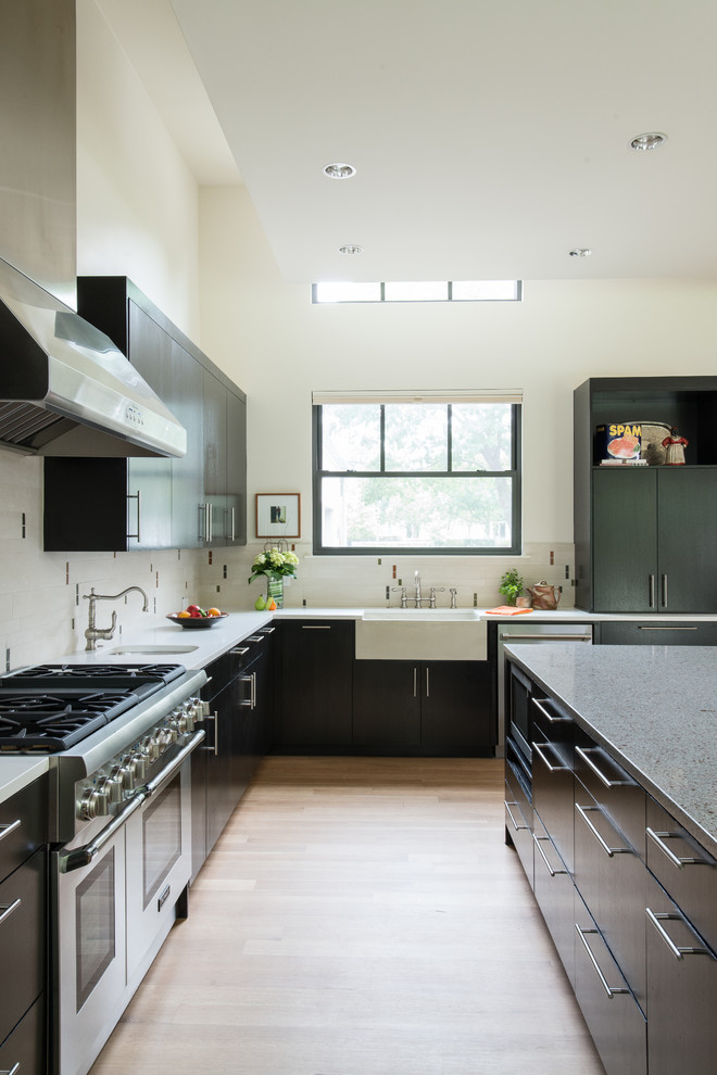 Inspiration for a mid-sized contemporary l-shaped open plan kitchen in Dallas with a farmhouse sink, flat-panel cabinets, black cabinets, quartz benchtops, white splashback, porcelain splashback, stainless steel appliances, light hardwood floors and with island.