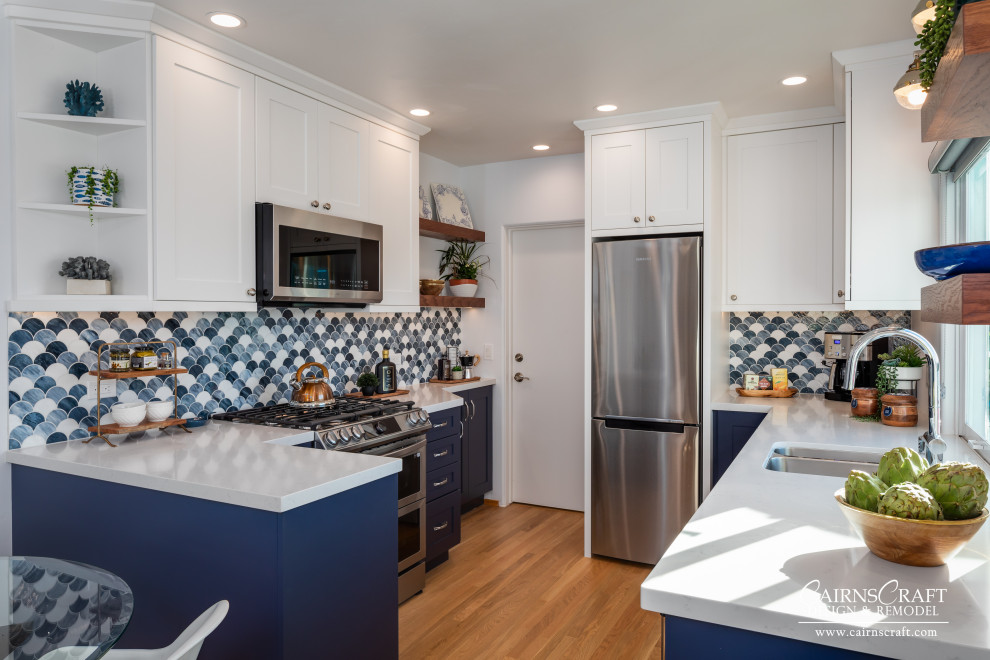Photo of a medium sized coastal u-shaped kitchen/diner in San Diego with a double-bowl sink, shaker cabinets, white cabinets, engineered stone countertops, blue splashback, glass sheet splashback, stainless steel appliances, medium hardwood flooring, a breakfast bar, brown floors and white worktops.